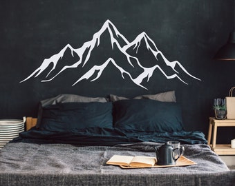 May include: A bedroom with a black wall featuring a white mountain range decal. The bed has a gray duvet and dark blue pillows. A wooden nightstand with a small potted plant and candles is on the right side of the bed.