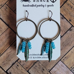May include: A pair of hoop earrings with turquoise and green feather charms. The earrings are on a white card with the text "krabbe shack handcrafted jewelry & goods" and a crab and anchor logo.