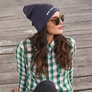 Puede incluir: Una mujer con una boina azul oscuro con el texto "your text here" bordado, una camisa de cuadros verde y blanco y unos vaqueros azules oscuros. Lleva gafas de sol marrones y mira hacia la derecha.