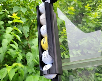 May include: A black golf ball holder with six golf balls inside. The holder is attached to a black metal pole. The golf balls are white and yellow.