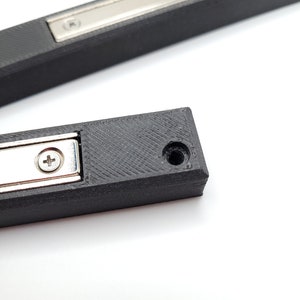 Peut inclure: Gros plan sur un loquet de porte magn&eacute;tique en plastique noir avec une plaque en m&eacute;tal argent&eacute; et un petit trou de vis. Le loquet est con&ccedil;u pour maintenir une porte ferm&eacute;e.