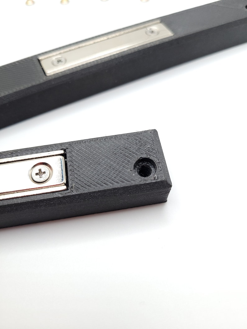 Peut inclure: Gros plan sur un loquet de porte magn&eacute;tique en plastique noir avec une plaque en m&eacute;tal argent&eacute; et un petit trou de vis. Le loquet est con&ccedil;u pour maintenir une porte ferm&eacute;e.