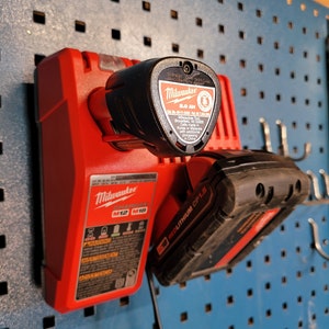 Peut inclure: Un chargeur de batterie d'outil &eacute;lectrique Milwaukee rouge avec une batterie d'outil &eacute;lectrique noire attach&eacute;e. Le chargeur porte l'inscription "Milwaukee M12 M18" et "REDLITHIUM CP1.5". La batterie porte l'inscription "Milwaukee 2.0 AH".