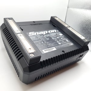 Puede incluir: Cargador de batería Snap-on CTC131A negro con diseño acanalado y detalles plateados. El cargador tiene una etiqueta con especificaciones técnicas y el logotipo de Snap-on. Tiene dos soportes rectangulares negros con placas metálicas plateadas.