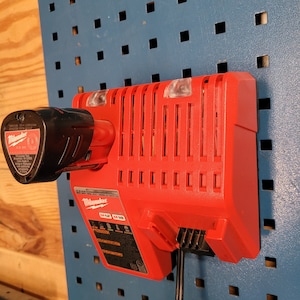 Peut inclure: Un chargeur Milwaukee M12 M18 rouge avec un cordon d'alimentation noir branch&eacute;. Le chargeur est mont&eacute; sur un panneau perfor&eacute; bleu.