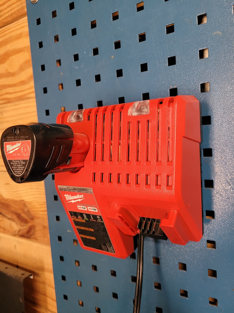 Peut inclure: Un chargeur Milwaukee M12 M18 rouge avec un cordon d'alimentation noir branch&eacute;. Le chargeur est mont&eacute; sur un panneau perfor&eacute; bleu.