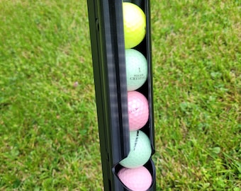 May include: A black golf ball holder with five golf balls inside. The golf balls are green, pink, and yellow.