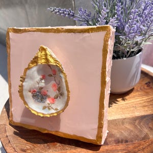 May include: A pink square with gold trim, featuring a white oyster shell with a floral design. The shell is painted with pink and white flowers and green leaves. The square is sitting on a wooden surface.