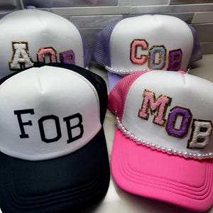 May include: Four trucker caps with white front panels and mesh backs. The caps display the letters "AOB", "COB", "FOB", and "MOB". Each cap has a different coloured brim and lettering, with some featuring pearl detailing.