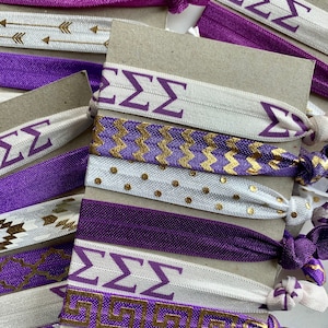 May include: A set of purple and white hair ties with gold accents. The hair ties feature different patterns, including chevron, polka dots, and Greek key designs. The hair ties also have the letters "ZZZ" printed on them.