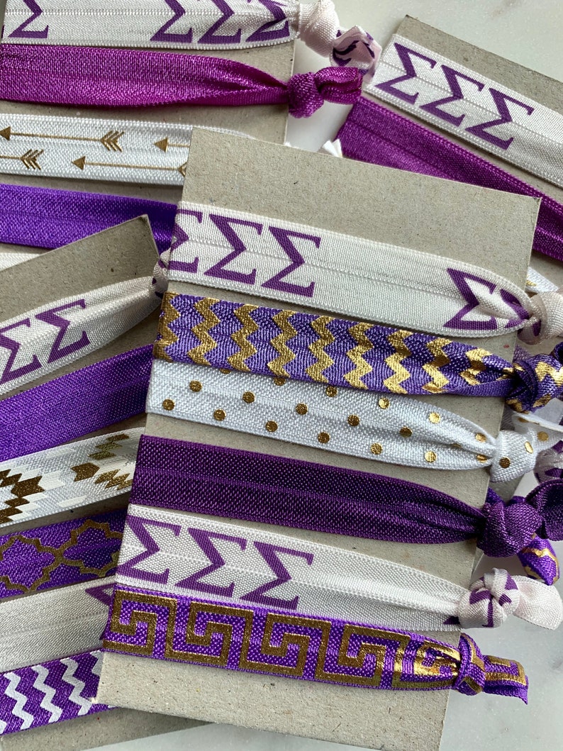 May include: A set of purple and white hair ties with gold accents. The hair ties feature different patterns, including chevron, polka dots, and Greek key designs. The hair ties also have the letters "ZZZ" printed on them.