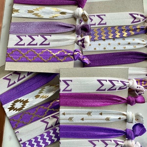 May include: A set of purple and white hair ties with gold accents. The hair ties feature a variety of patterns, including chevron, polka dots, and geometric designs. The hair ties also have the letters "&Sigma;&Sigma;&Sigma;" printed on them.