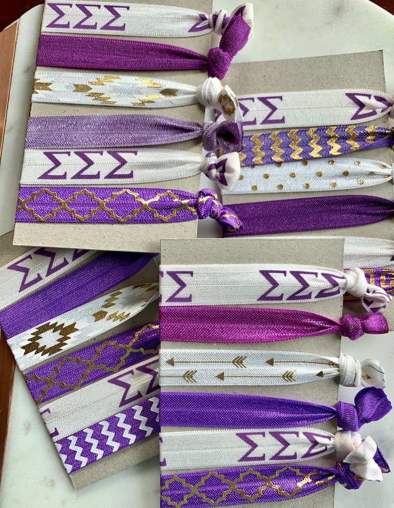 May include: A set of purple and white hair ties with gold accents. The hair ties feature a variety of patterns, including chevron, polka dots, and geometric designs. The hair ties also have the letters "&Sigma;&Sigma;&Sigma;" printed on them.