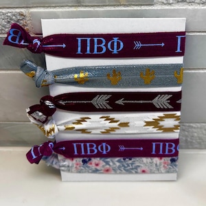 May include: A set of six hair ties in various colors and patterns. The ties include burgundy, gray, and white bands with gold and blue accents. Some bands feature text and arrow designs. The ties are displayed on a white card.