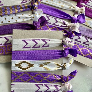 May include: A set of purple and white hair ties with gold accents. The hair ties feature different patterns, including chevron, polka dots, and Greek letters. The hair ties are arranged on a brown surface.