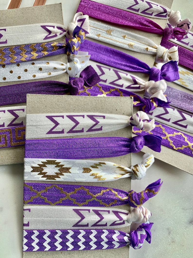 May include: A set of purple and white hair ties with gold accents. The hair ties feature different patterns, including chevron, polka dots, and Greek letters. The hair ties are arranged on a brown surface.