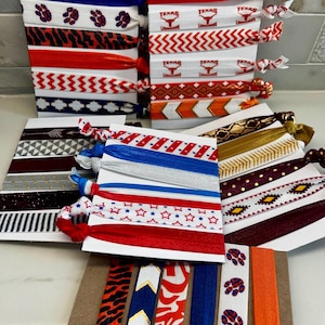 May include: A selection of hair ties in diverse colours and patterns. Designs include paw prints, zigzags, stars, and the word "Texas" with a longhorn logo. The hair ties are displayed on white and brown card.