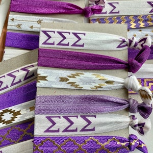 May include: A set of purple and white hair ties with gold accents. The hair ties feature different patterns, including arrows, zigzags, and geometric designs. The hair ties also have the letters "ZZZ" printed on them.