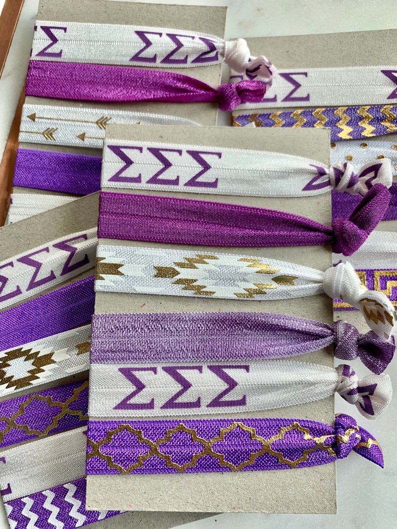 May include: A set of purple and white hair ties with gold accents. The hair ties feature different patterns, including arrows, zigzags, and geometric designs. The hair ties also have the letters "ZZZ" printed on them.