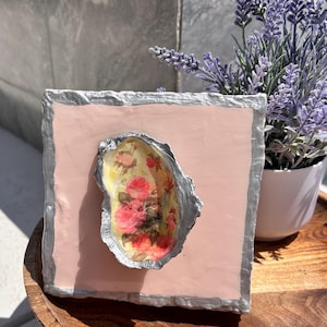 May include: A pink square with a silver border, featuring a decorative oyster shell with a floral design. The shell is centred on the square and has a light yellow background with pink and yellow flowers.