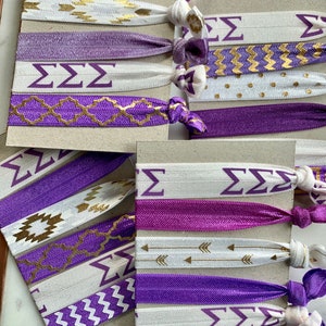 May include: A set of purple and white hair ties with gold designs. The hair ties feature different patterns, including geometric shapes, arrows, and the Greek letters "&Sigma;&Sigma;&Sigma;".