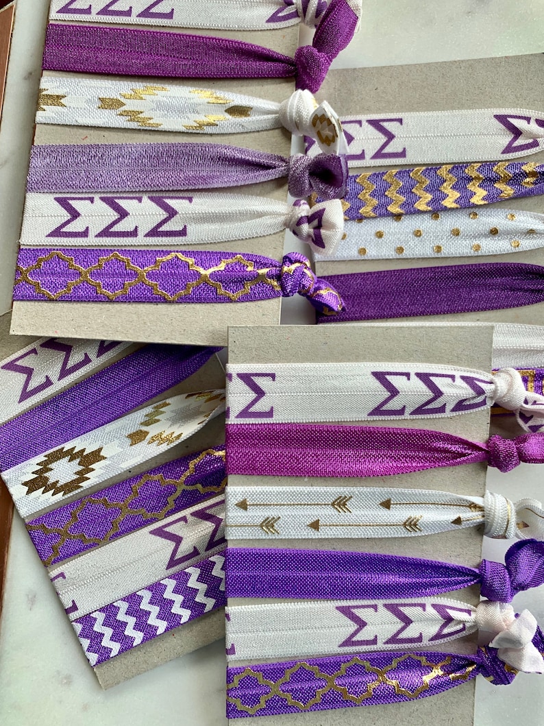 May include: A set of purple and white hair ties with gold designs. The hair ties feature different patterns, including geometric shapes, arrows, and the Greek letters "&Sigma;&Sigma;&Sigma;".