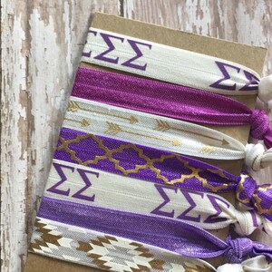 May include: A set of six purple and white hair ties with gold accents. The hair ties feature different patterns, including arrows, geometric shapes, and the Greek letters "&Sigma;&Sigma;".