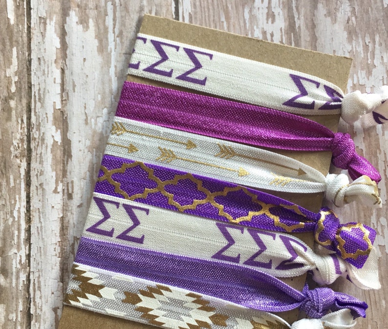 May include: A set of six purple and white hair ties with gold accents. The hair ties feature different patterns, including arrows, geometric shapes, and the Greek letters "&Sigma;&Sigma;".