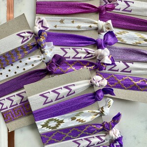 May include: A set of purple and white hair ties with gold accents. The hair ties feature different patterns, including chevron, polka dots, and geometric designs. The hair ties also have the Greek letters "&Sigma;&Sigma;&Sigma;" printed on them.