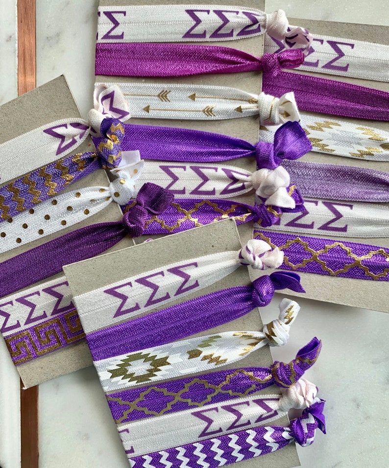 May include: A set of purple and white hair ties with gold accents. The hair ties feature different patterns, including chevron, polka dots, and geometric designs. The hair ties also have the Greek letters "&Sigma;&Sigma;&Sigma;" printed on them.