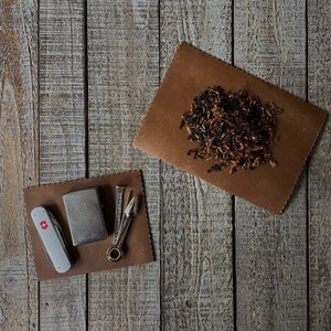 Puede incluir: Una bolsa de cuero marrón con un encendedor plateado y una pequeña herramienta plateada con una punta dorada. La bolsa está llena de tabaco suelto.