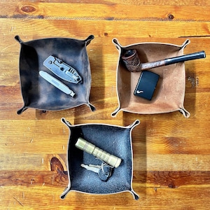 May include: Three leather catchall trays in varying colors. One tray holds a pocket knife and a small pen. Another holds a pipe and a black lighter. The third tray contains a flashlight and keys. The trays are square with rounded corners.
