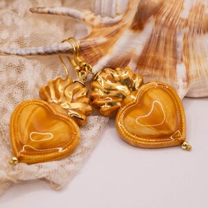 May include: A pair of gold-colored heart-shaped earrings with a glossy finish. The earrings have a textured, ornate design and are suspended from gold-colored hooks. The earrings are displayed on a cream-colored lace fabric, with a seashell in the background.