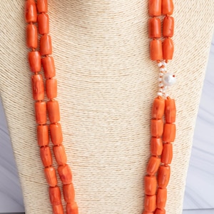 May include: A double-strand necklace featuring vibrant orange barrel-shaped beads. The necklace is accented with small white beads and a single, larger white bead. The jewelry is displayed on a cream-colored mannequin.