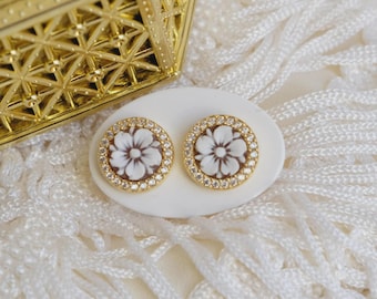 May include: A pair of gold-toned stud earrings with a floral cameo design. Each earring features a white flower on a brown background, surrounded by a ring of clear crystals. The earrings are displayed on a white oval surface.