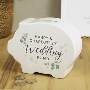 May include: A white ceramic piggy bank shaped like a pig, with the text "HARRY & CHARLOTTE'S Wedding FUND" in black script. The design includes gray hearts and green and gray eucalyptus leaves. Coins are visible near the bank.