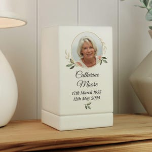 May include: White ceramic memorial candle holder with a photo of a woman, green leaves, and the text "Catherine Moore, 17th March 1955, 12th May 2025".