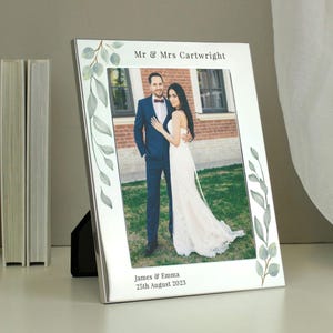 May include: Silver photo frame with a white mount and a leafy border. The frame has a photo of a couple in their wedding attire. The text on the frame reads "Mr & Mrs Cartwright, James & Emma, 25th August 2023".