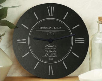 May include: A round black slate wall clock featuring white Roman numerals and two black hands. The center is engraved with the names SIMON AND KELLY and the text Time is not measured by Clocks but by Moments, surrounded by decorative line accents.