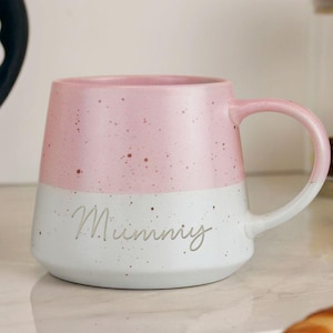 May include: A two-tone ceramic mug with a pink speckled top and a white speckled bottom. The word "Mummy" is written in cursive on the white section. The mug has a curved handle.