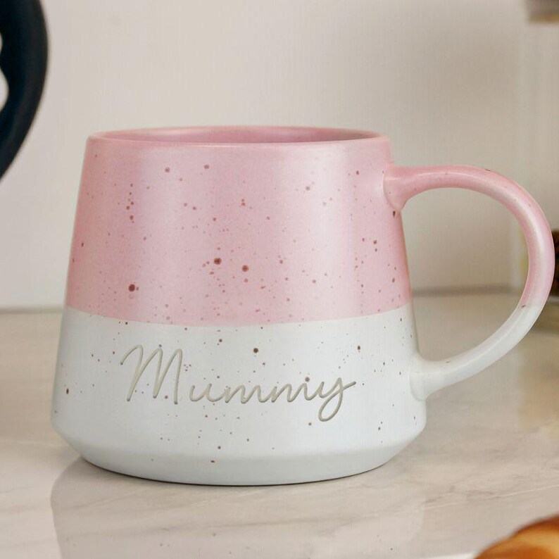 May include: A two-tone ceramic mug with a pink speckled top and a white speckled bottom. The word "Mummy" is written in cursive on the white section. The mug has a curved handle.