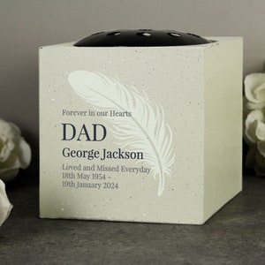 May include: A square, pale-coloured memorial flower vase with a black top. The vase has a feather design and the text "Forever in our Hearts," "DAD," the name "George Jackson," and dates.