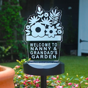 May include: White solar-powered garden sign with a flower pot design and the text "Welcome to Nanny & Grandad's Garden".