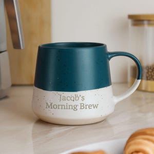 May include: A teal and white ceramic mug with the text "Jacob's Morning Brew." The mug has a speckled white base and a teal upper half. The handle is white with a teal accent. A jar and wooden cutting board are in the background.