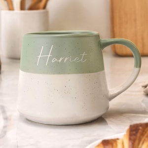 May include: A two-tone ceramic mug with a mint green top and white bottom. The word "Harriet" is written in cursive on the front. The mug has a rounded handle and a speckled texture.
