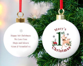 May include: Two white spherical Christmas ornaments with gold caps and red ribbons. One features a fawn illustration, a mushroom, and the text Macy's 1st Christmas. The other displays the text Happy 1st Christmas We Love You Hugs and Kisses Gran & Grandad xx.