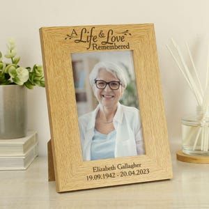 May include: A wooden photo frame with a light brown finish. The frame has a white mount and a clear glass front. The frame is engraved with the text "A Life & Love Remembered". The frame also includes a personalised inscription with a name and dates.