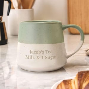 May include: A two-tone ceramic mug with a mint green top and white bottom. The mug has a rounded handle and the text "Jacob's Tea Milk & 1 Sugar" is printed on the front. The mug is on a marble surface.