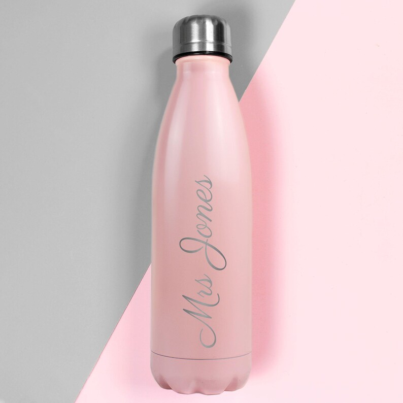 May include: A pink insulated water bottle with a silver cap. The bottle has the text "Mrs Jones" in a cursive font. The background is split diagonally, with gray on the left and pink on the right.