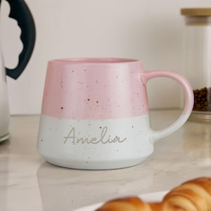 May include: A two-tone ceramic mug with a pink upper half and a white lower half, speckled with brown dots. The name "Amelia" is written in cursive on the white section. The mug has a curved handle.